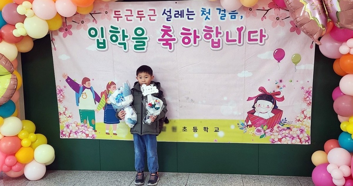 ‘두근두근 설레는 첫걸음, 입학을 축하합니다’ 현수막과 풍선 장식 앞에 꽃다발을 안고 선 아이