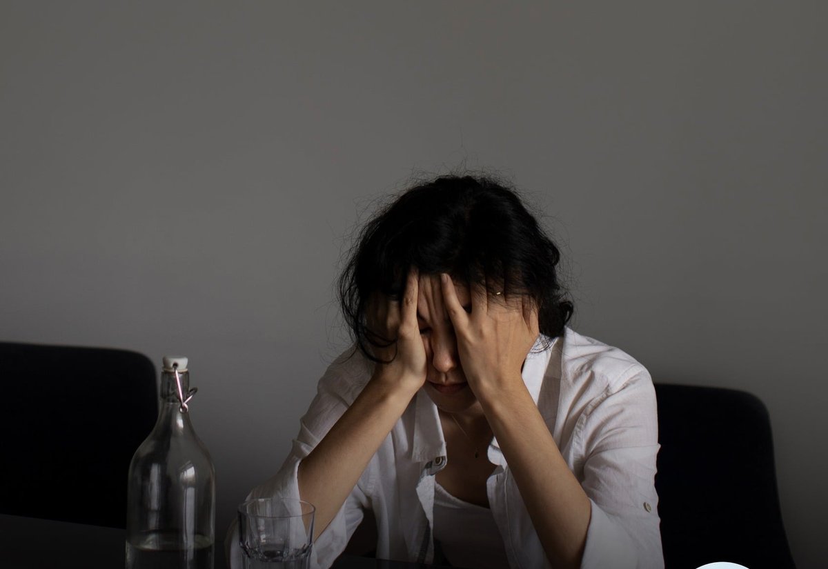 어두운 탁자에 앉아 두 손으로 이마를 감싼 여성과 물병·유리잔