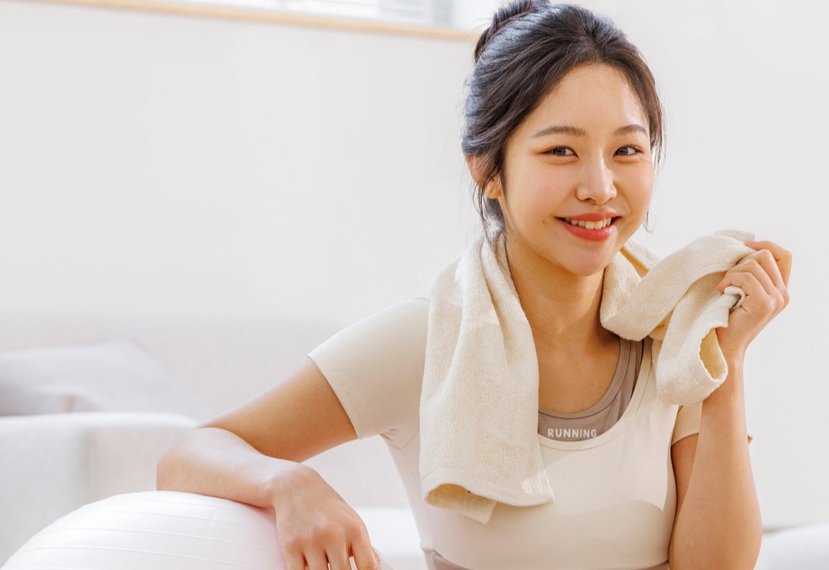 목에 수건을 두르고 짐볼에 팔을 기댄 채 웃는 운동복 차림의 여성