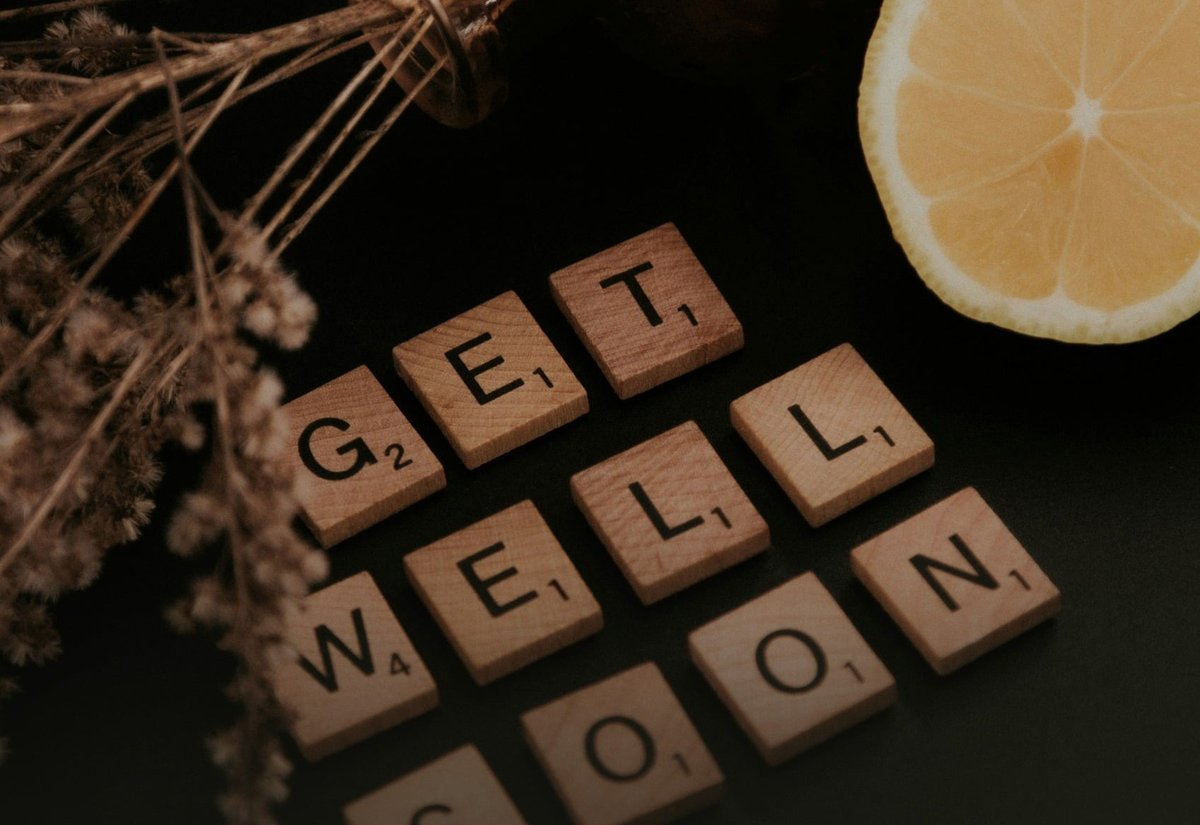 어두운 바탕 위 갈색 유리병과 마른 꽃, 반으로 자른 레몬, 'GET WELL SOON' 글자 나무 타일