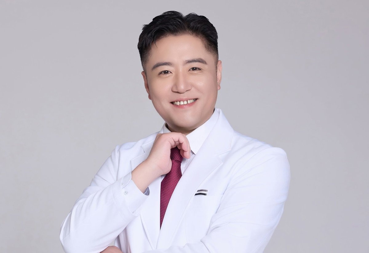 흰 가운에 붉은 넥타이를 매고 웃으며 팔짱을 낀 남성
