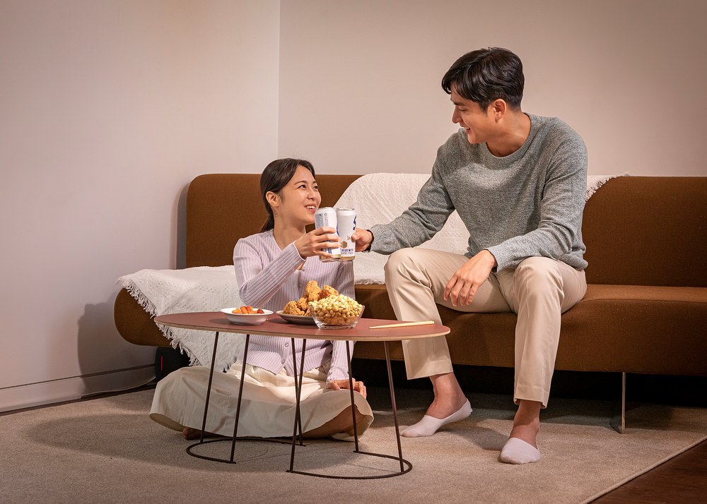 거실 소파 앞 테이블에 간식을 두고 캔 음료로 건배하는 남녀